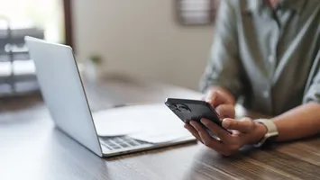 A person's hands using their mobile phone while sitting in front of a laptop